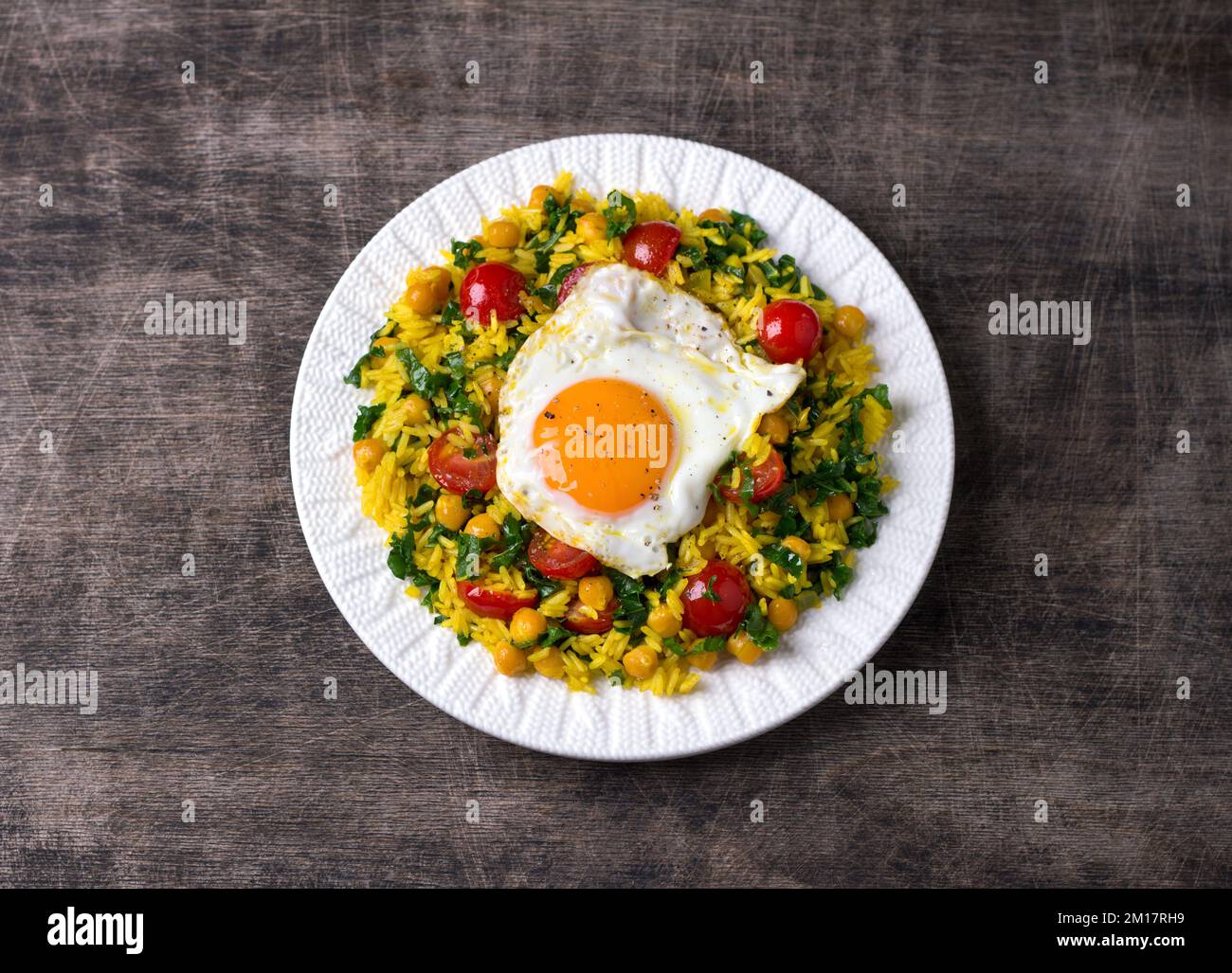 Chinese fried rice with turmeric, chickpeas, tomatoes, kale and with fried eggs on white ceramic plate on dark wooden background. Quick and easy dinne Stock Photo