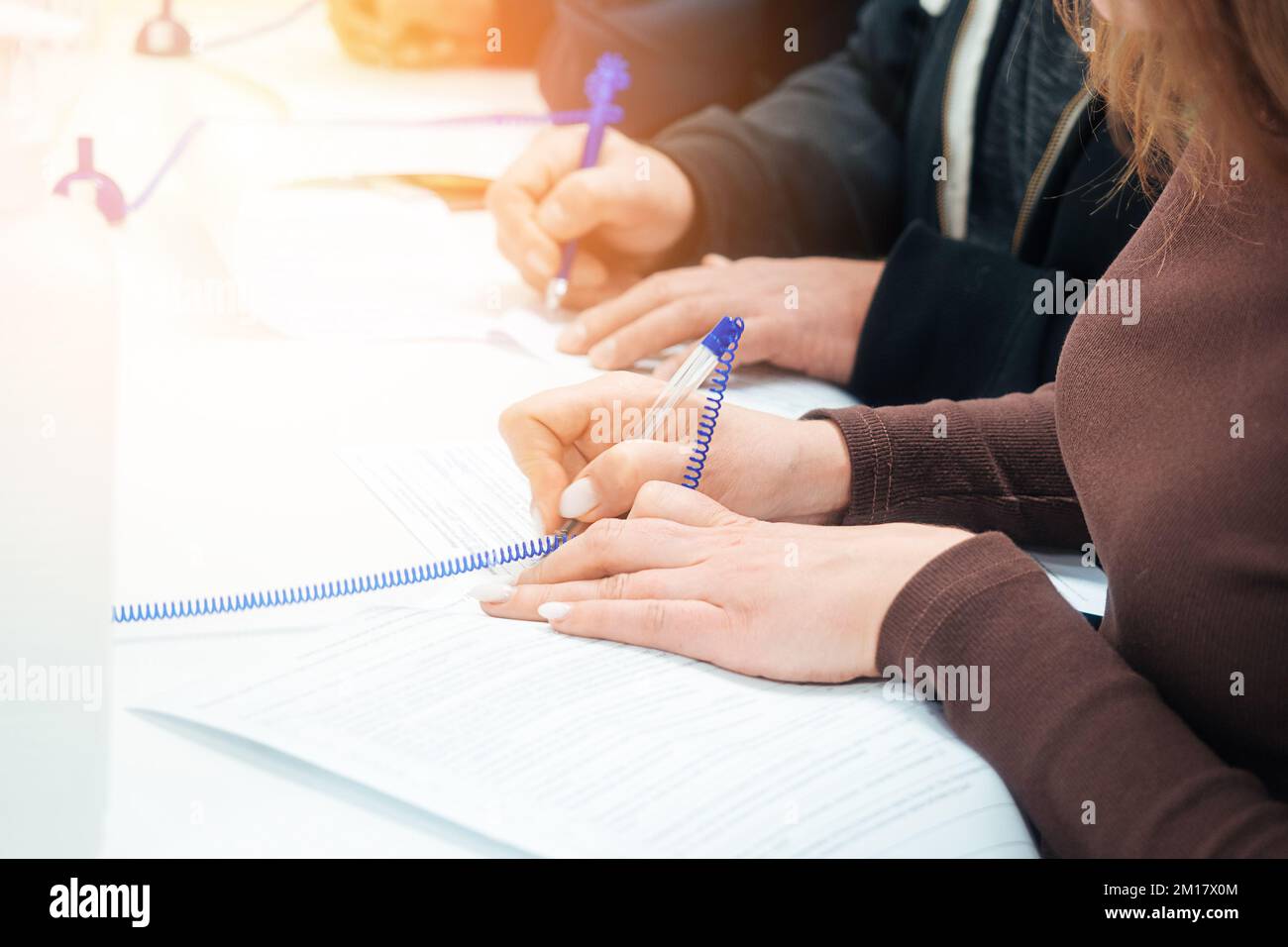 Woman fills out blank application form at table or signs contract. Hands of people with close-up fountain pen. Perspective view Several people simultaneously fill in data with pen on paper.. Stock Photo