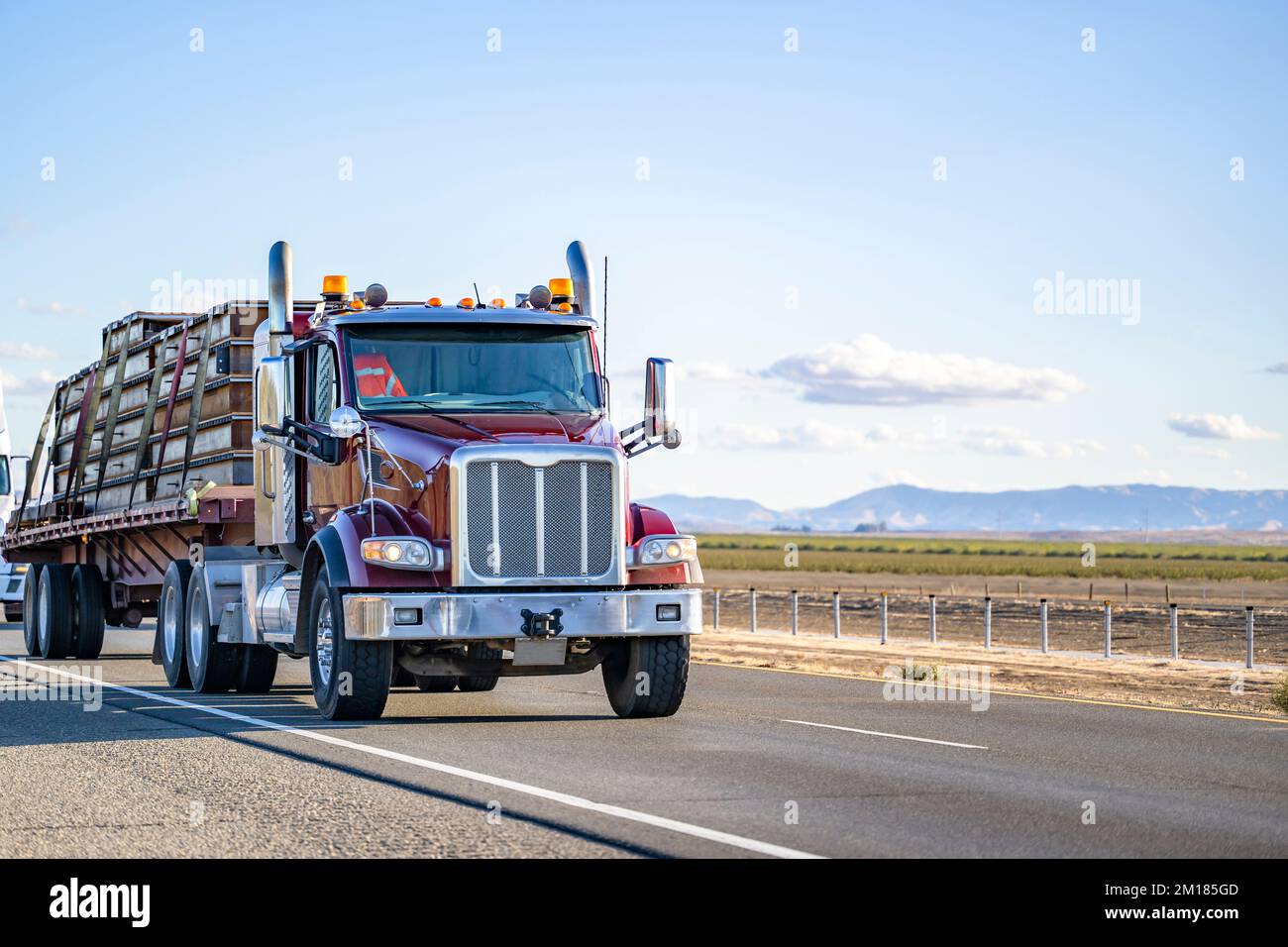 Industrial long hauler big rig dark red semi truck with chrome parts transporting fastened by slings commercial cargo on flat bed semi trailer running Stock Photo