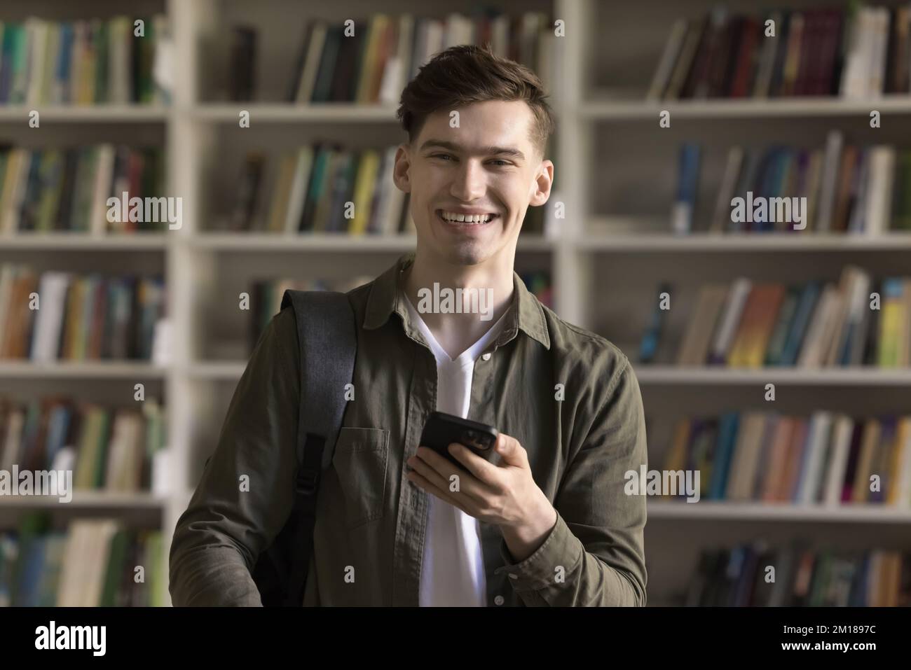 Happy handsome college student guy holding smartphone, posing in library Stock Photo