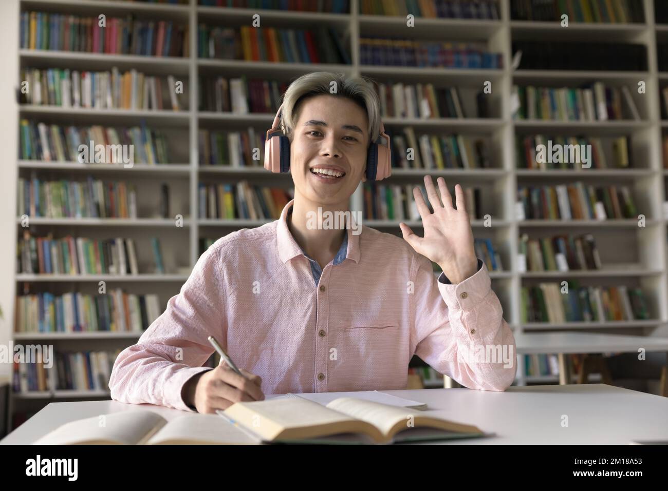 Happy Asian student guy in pink headphones waving hand hello Stock Photo