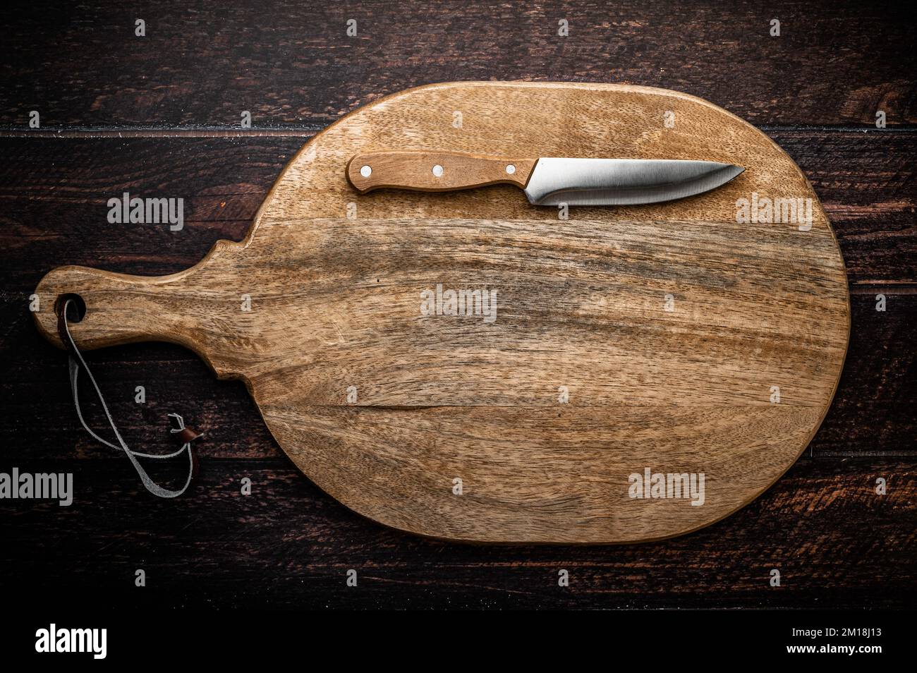 A large wooden cutting board with a knife on a dark wooden table. Stock Photo