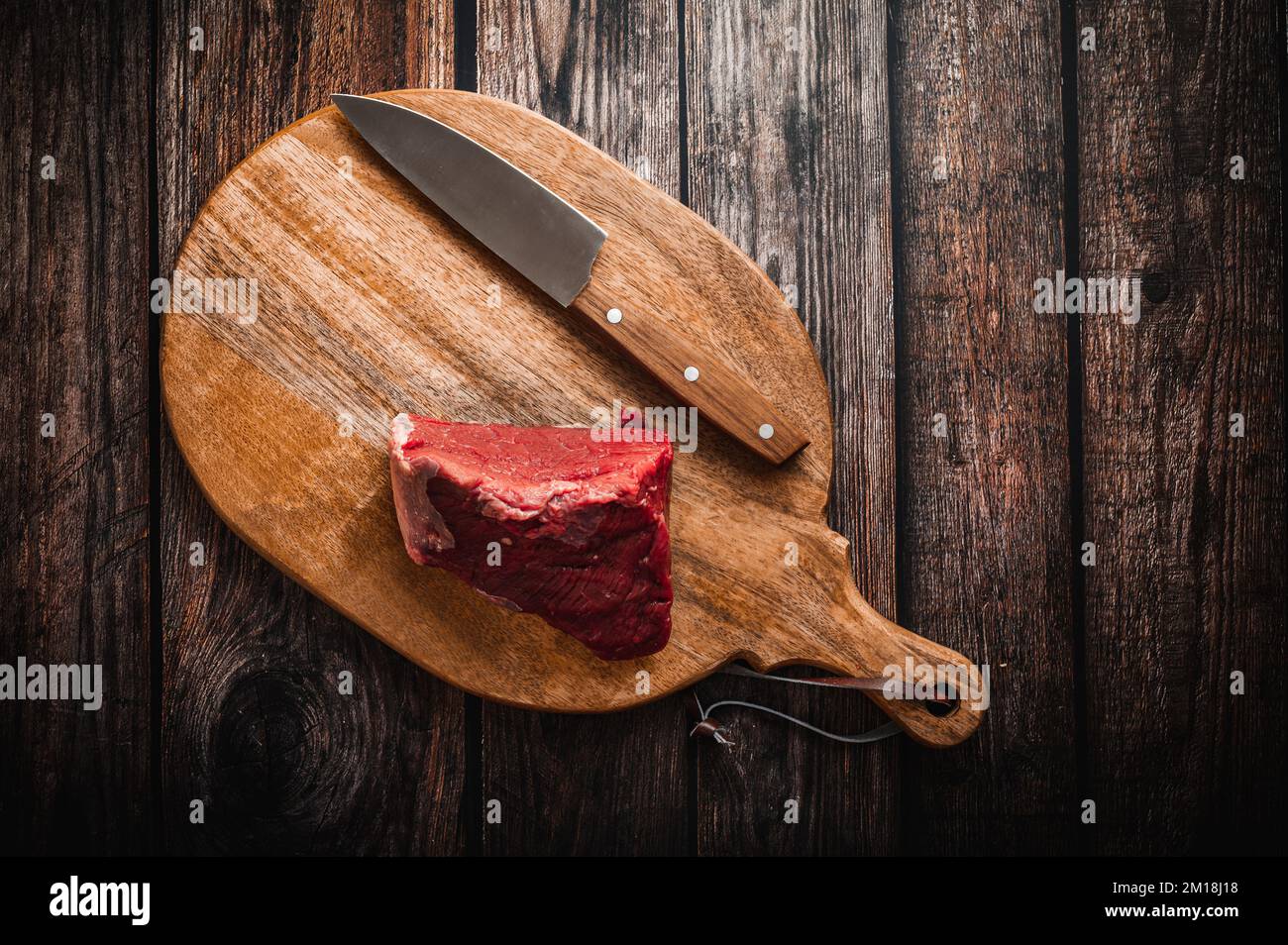 A piece of raw beef on a wooden cutting board with a knife. Wooden table, top down view, flat lay. Stock Photo
