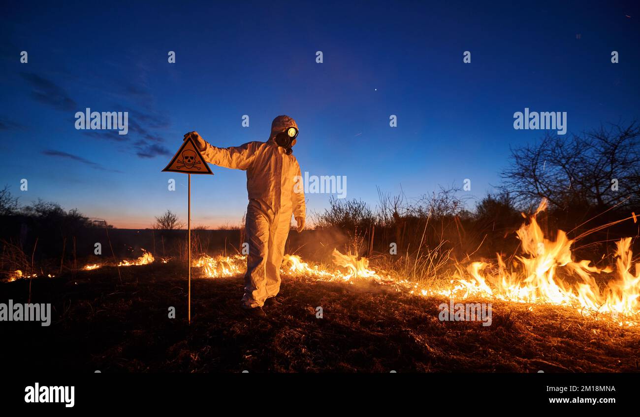 Fireman ecologist fighting fire in field at night. Man in protective suit and gas mask near burning grass with smoke, holding warning sign with skull and crossbones. Natural disaster concept. Stock Photo