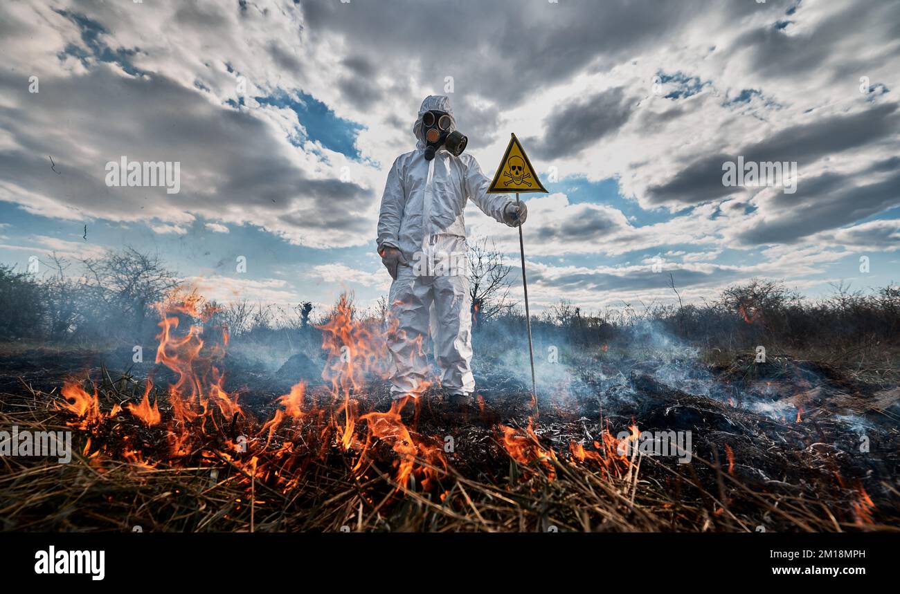 Firefighter ecologist fighting fire in field. Man in protective suit and gas mask near burning grass with smoke, holding warning sign with skull and crossbones. Natural disaster concept. Stock Photo
