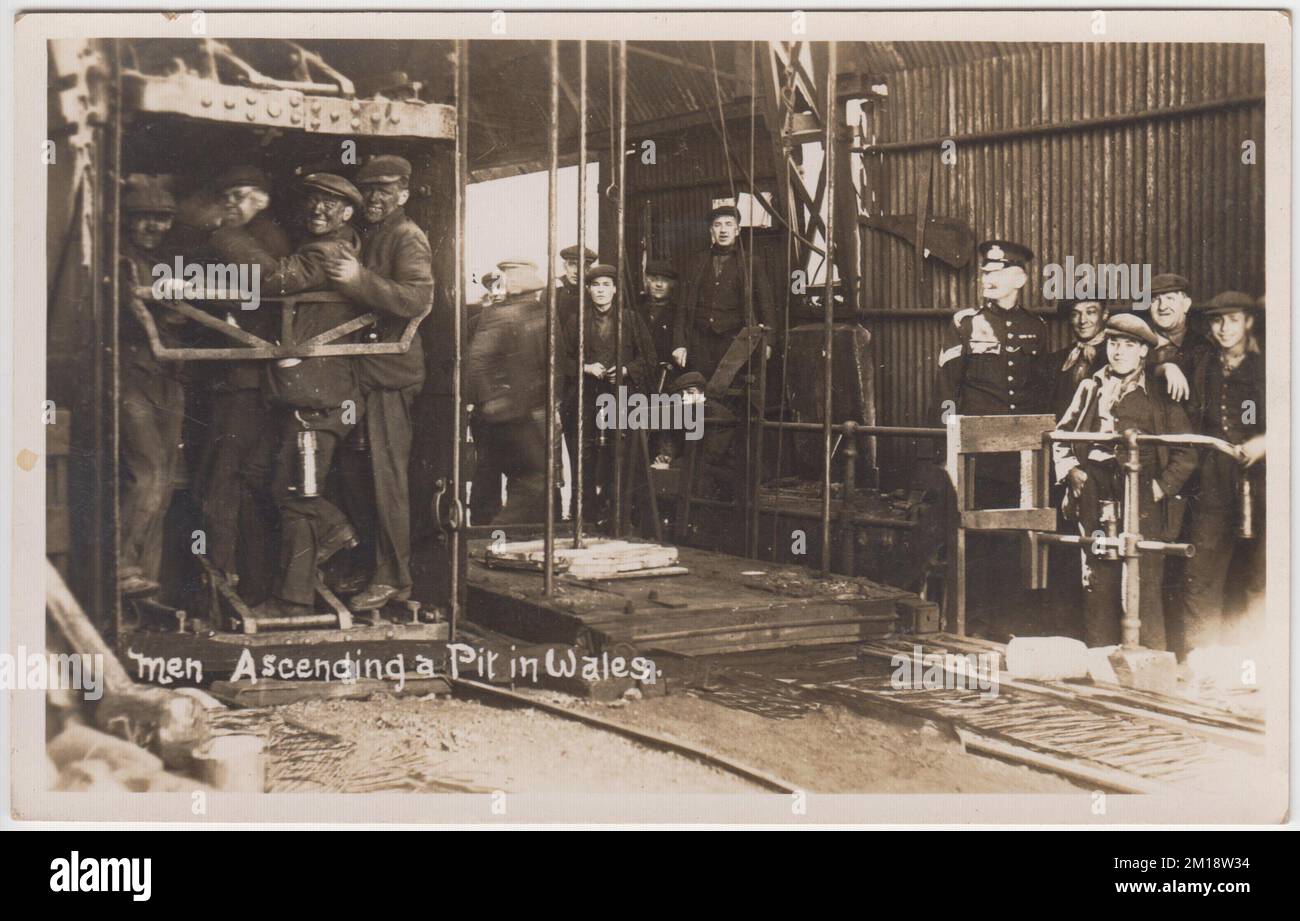 Coal mining - men ascending a pit in Wales. Early 20th century photograph of men with faces blackened by coal, crammed into the pit lift, coming up after the end of their shift. Men and boys are waiting in the background to go down the pit. Everyone is wearing a flat cap, with the exception of one man in uniform Stock Photo