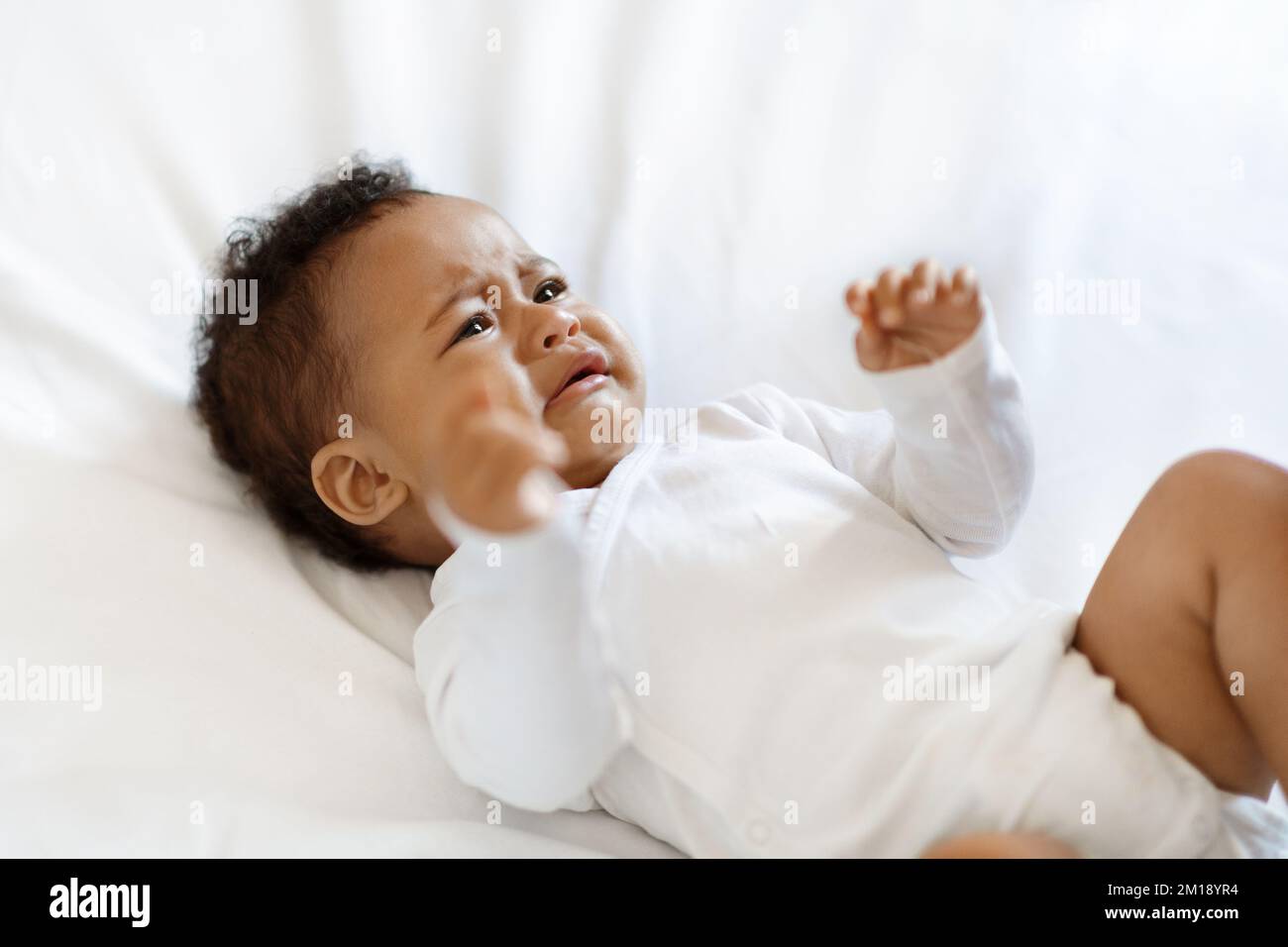 Portrait Of Crying Little Black Baby Wearing Bodysuit Lying On Bed Stock Photo