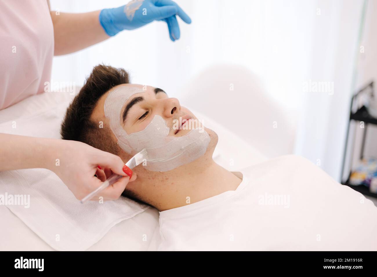 Cosmetologist applying clay mask on man's face in spa salon. Beautician make beuty for handsome man in beauty studio Stock Photo