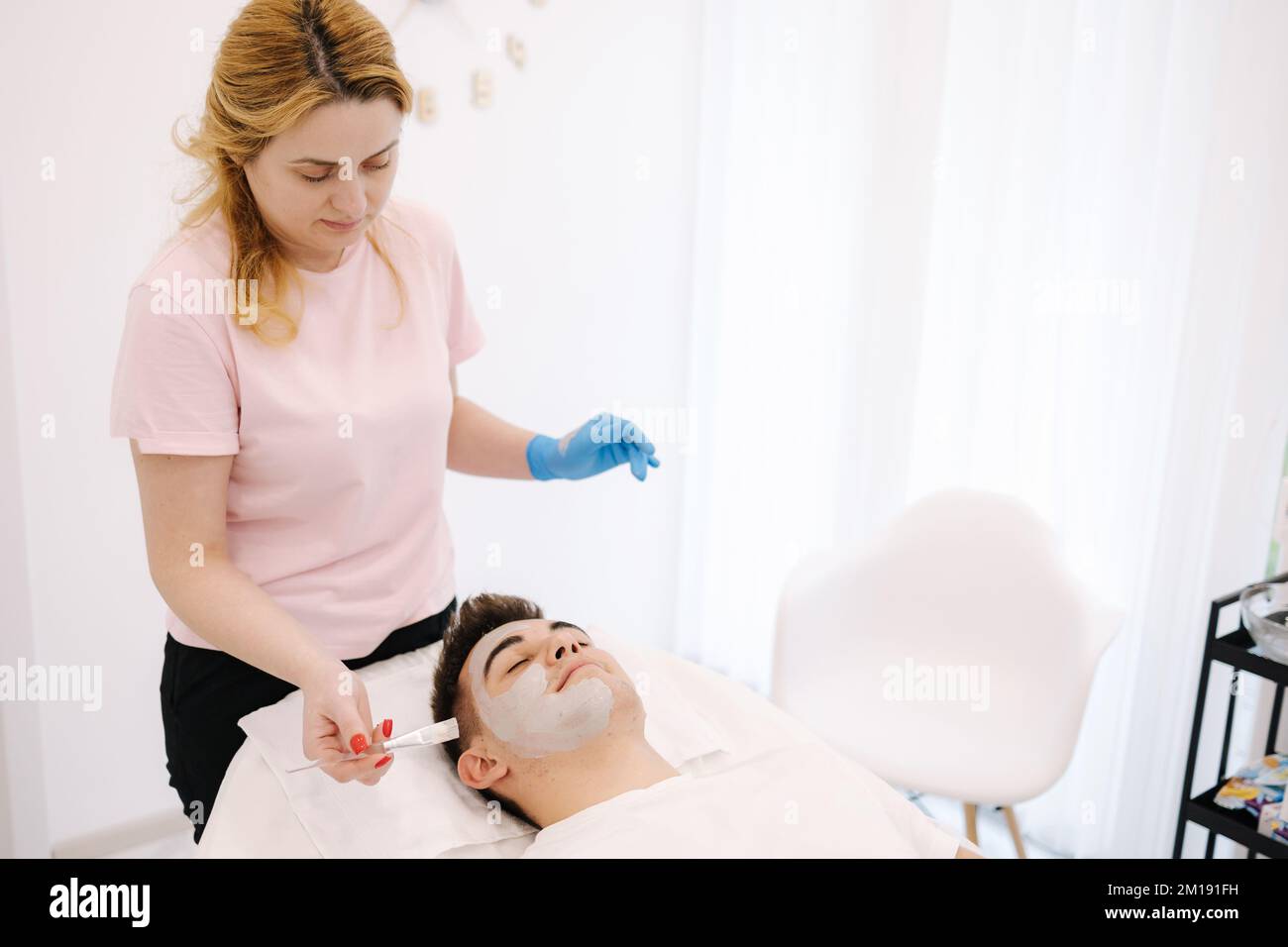 Cosmetologist applying clay mask on man's face in spa salon. Beautician make beuty for handsome man in beauty studio Stock Photo