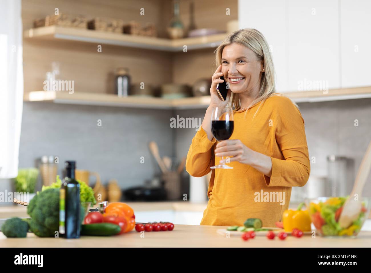 Happy blonde woman drinking wine while cooking, having phone