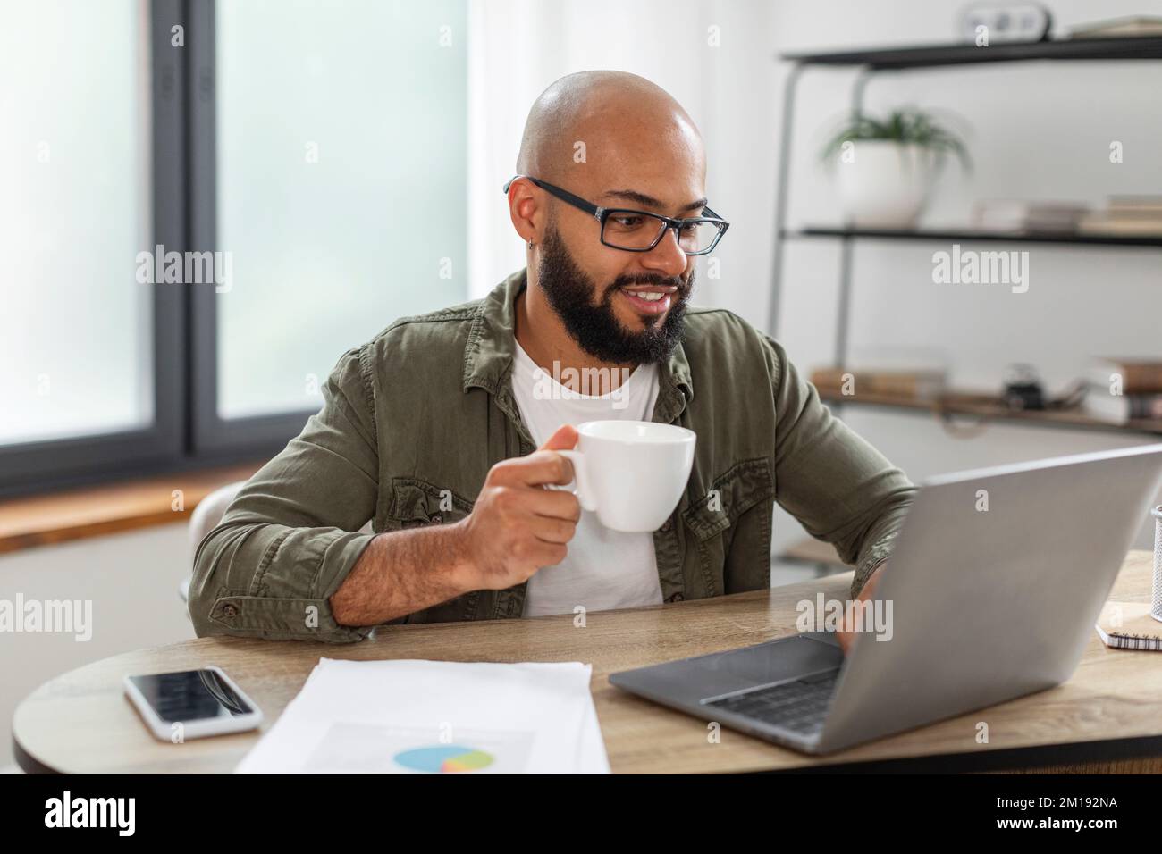 Positive latin bearded man using laptop, holding cup and enjoying drinking morning coffee while working from home Stock Photo