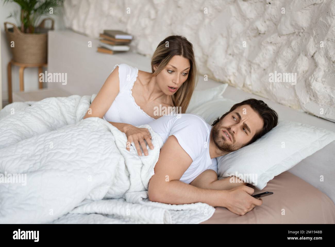 Shocked woman seeing her husband messaging to his lover on cellphone while lying in bed near him Stock Photo