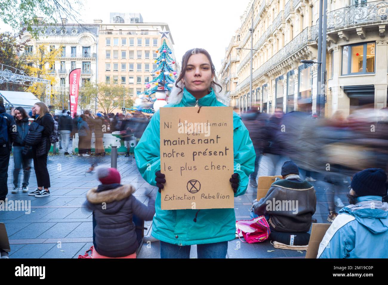 Toulouse, France. 11th Dec, 2022. A citizen standing in the middle of moving passers-by with a placard, Not yesterday, not tomorrow, now is our dearest present. XR Extinction Rebellion. Sitting / medit action, non-blocking, to cogitate and make cogitate on overconsumption during Christmas. in front of agitation and frenzy, calm and serenity; in front of massive consumption, happy sobriety; in front of injustices and social inequalities, solidarity and collaboration; in front of repression, gratitude to those who defend the living. France, Toulouse on December 10, 2022. Photo by Patricia Huchot Stock Photo