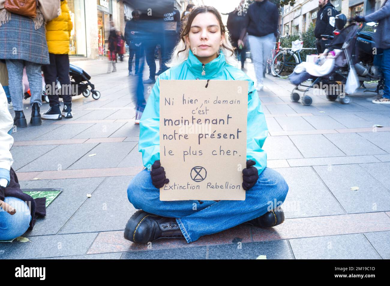 Toulouse, France. 11th Dec, 2022. A citizen sitting in the middle of the passers-by with a placard, Neither yesterday, nor tomorrow, it is now our dearest present. XR Extinction Rebellion. Sitting / medit action, non-blocking, to cogitate and make cogitate on overconsumption during Christmas. in front of agitation and frenzy, calm and serenity; in front of massive consumption, happy sobriety; in front of injustices and social inequalities, solidarity and collaboration; in front of repression, gratitude to those who defend the living. France, Toulouse on December 10, 2022. Photo by Patricia Huc Stock Photo