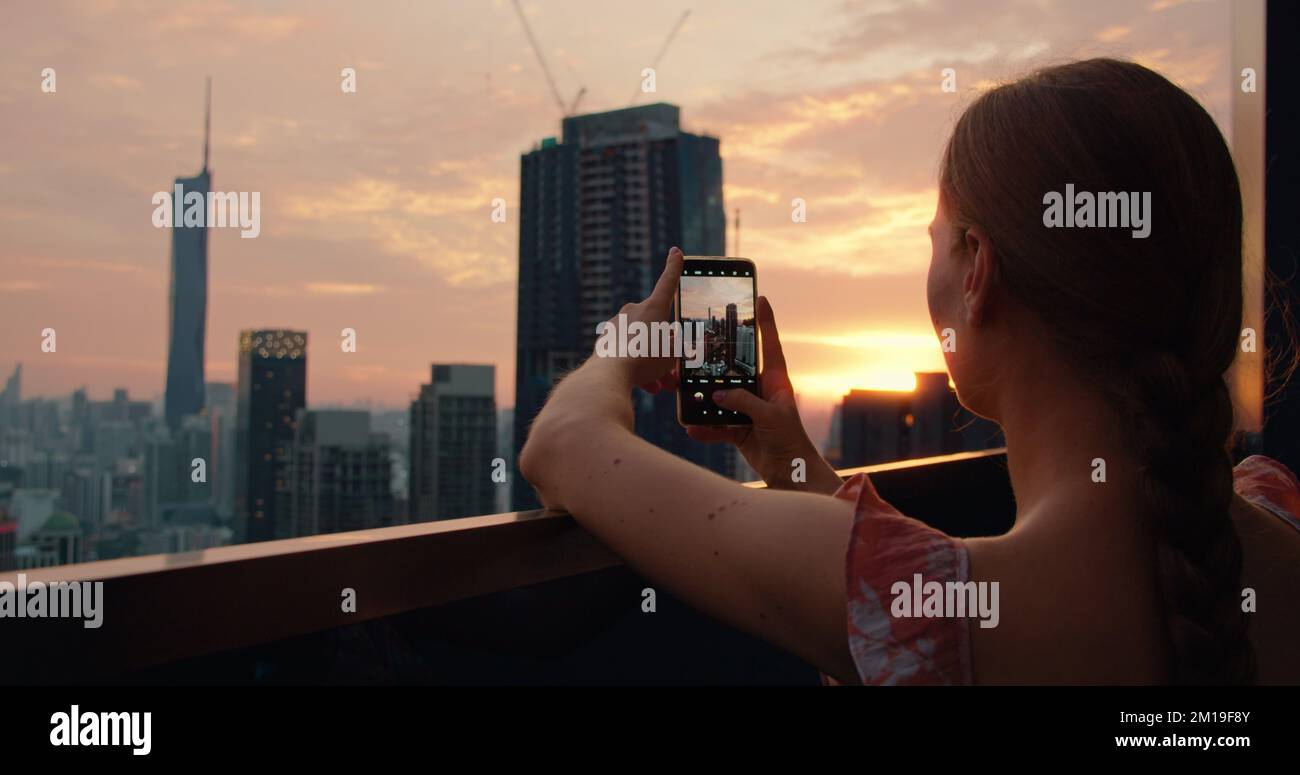Tourist woman taking photograph of cityscape skyscrapers at sunset in capital of Malaysia. Cloudy twilight peach skyline view from the roof of an apar Stock Photo