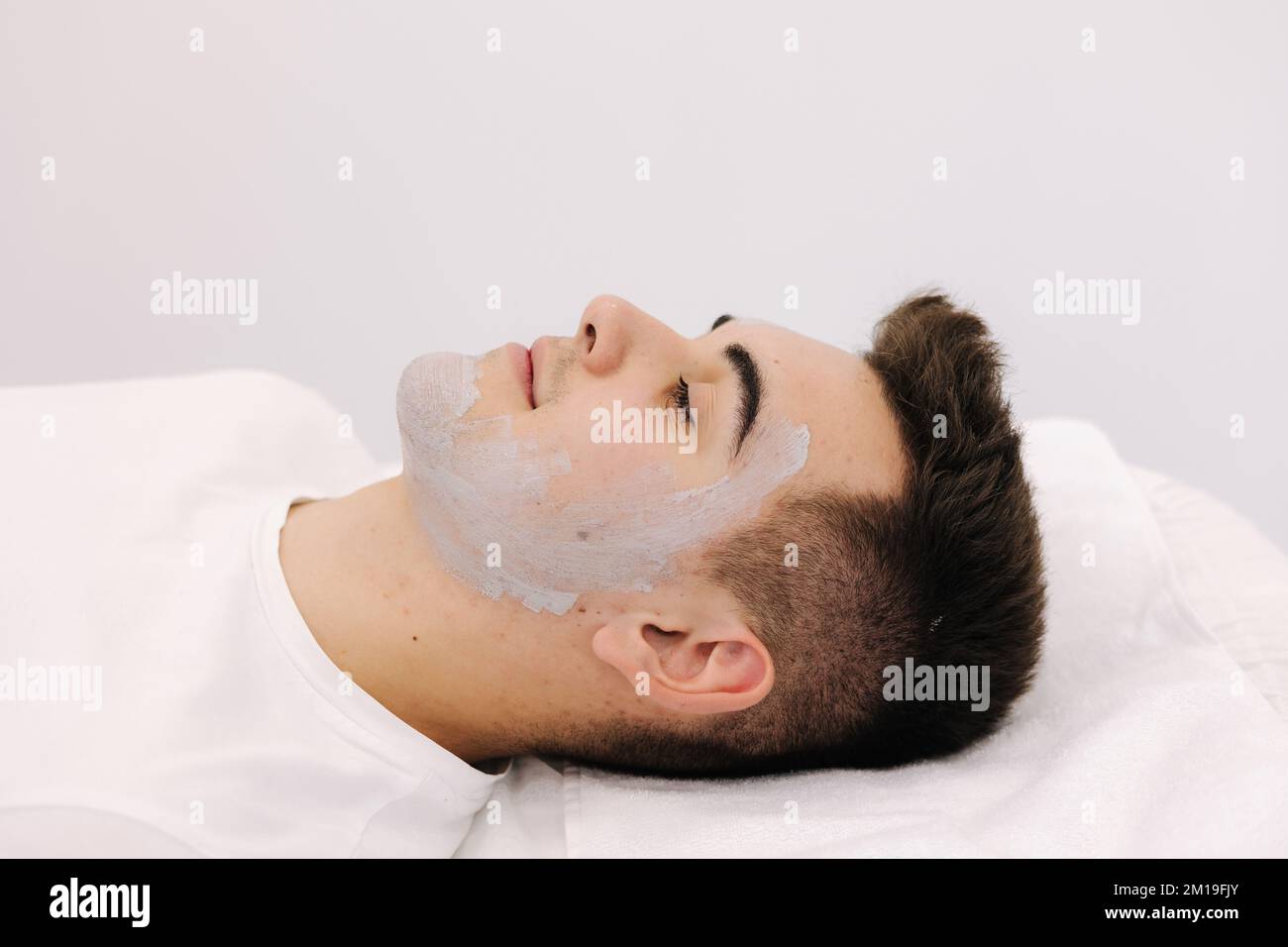 Cosmetologist applying clay mask on man's face in spa salon. Side view of male face during procedure. Stock Photo