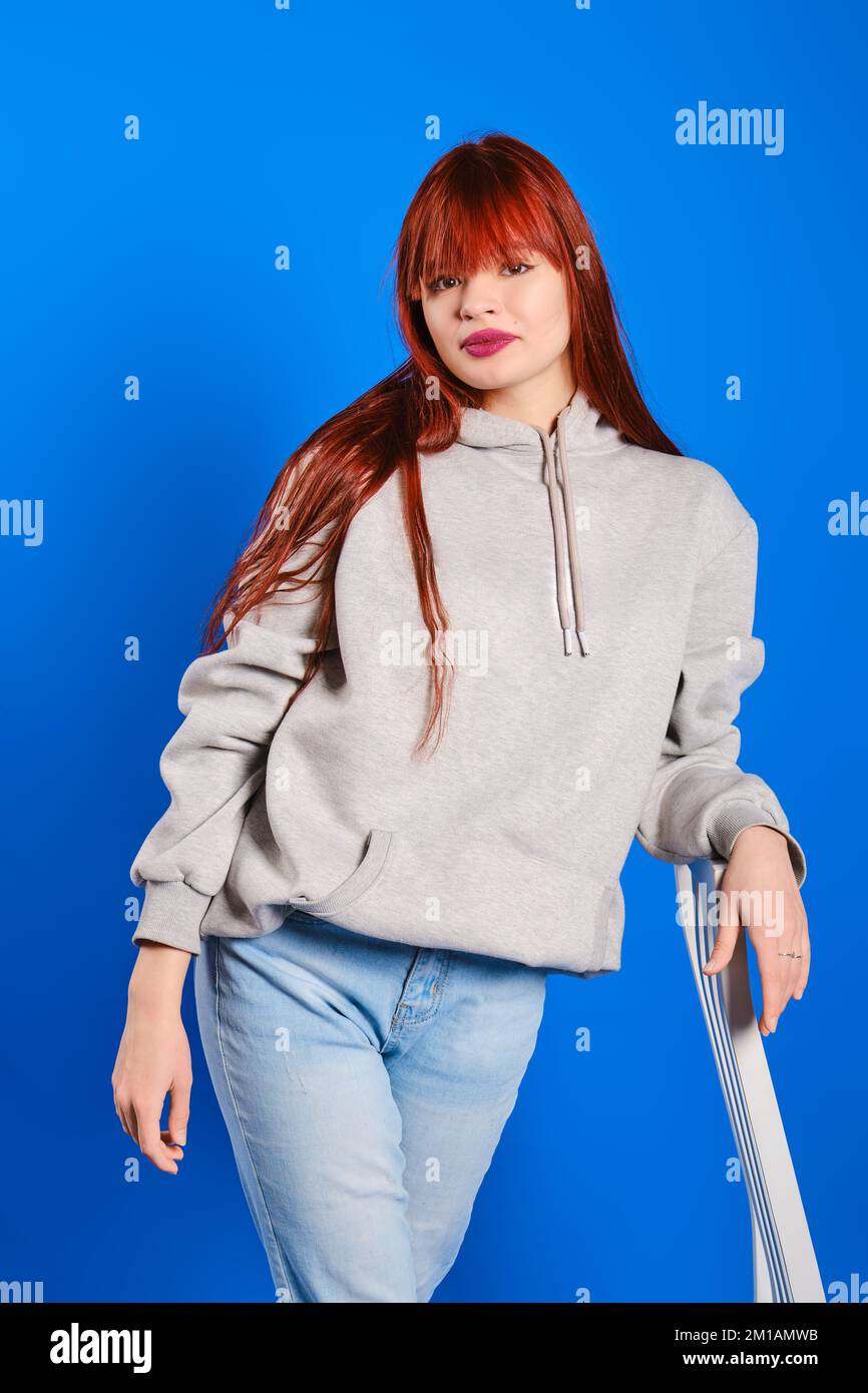 Studio portrait of young woman in blue jeans and grey hoodie on blue background Stock Photo