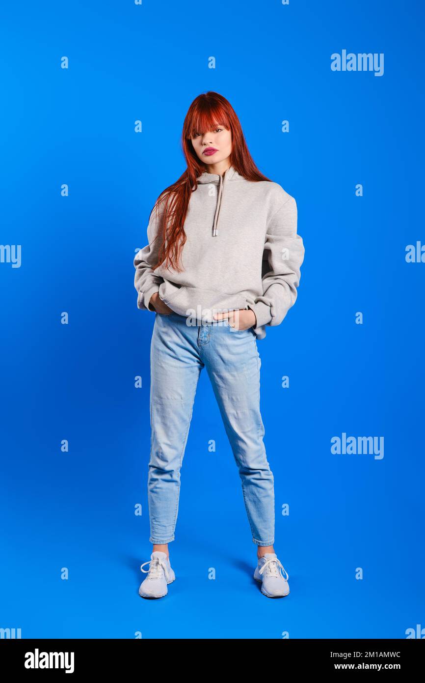Full length portrait of young red headed woman in blue jeans and grey hoodie on blue background Stock Photo
