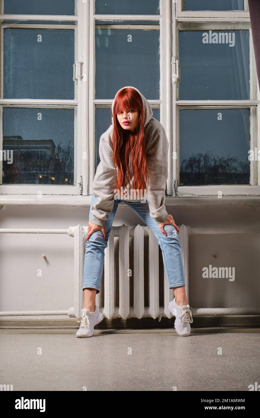 Casual portrait of young woman in jeans and sweatshirt with hood sitting on windowsill Stock Photo