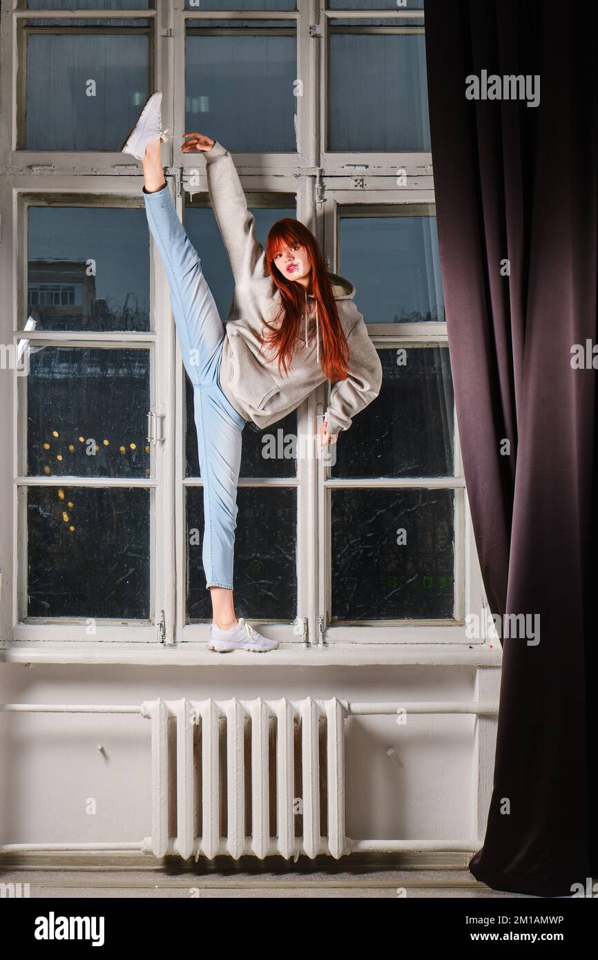 Young flexible woman in jeans and sweatshirt stretching leg while standing on windowsill Stock Photo
