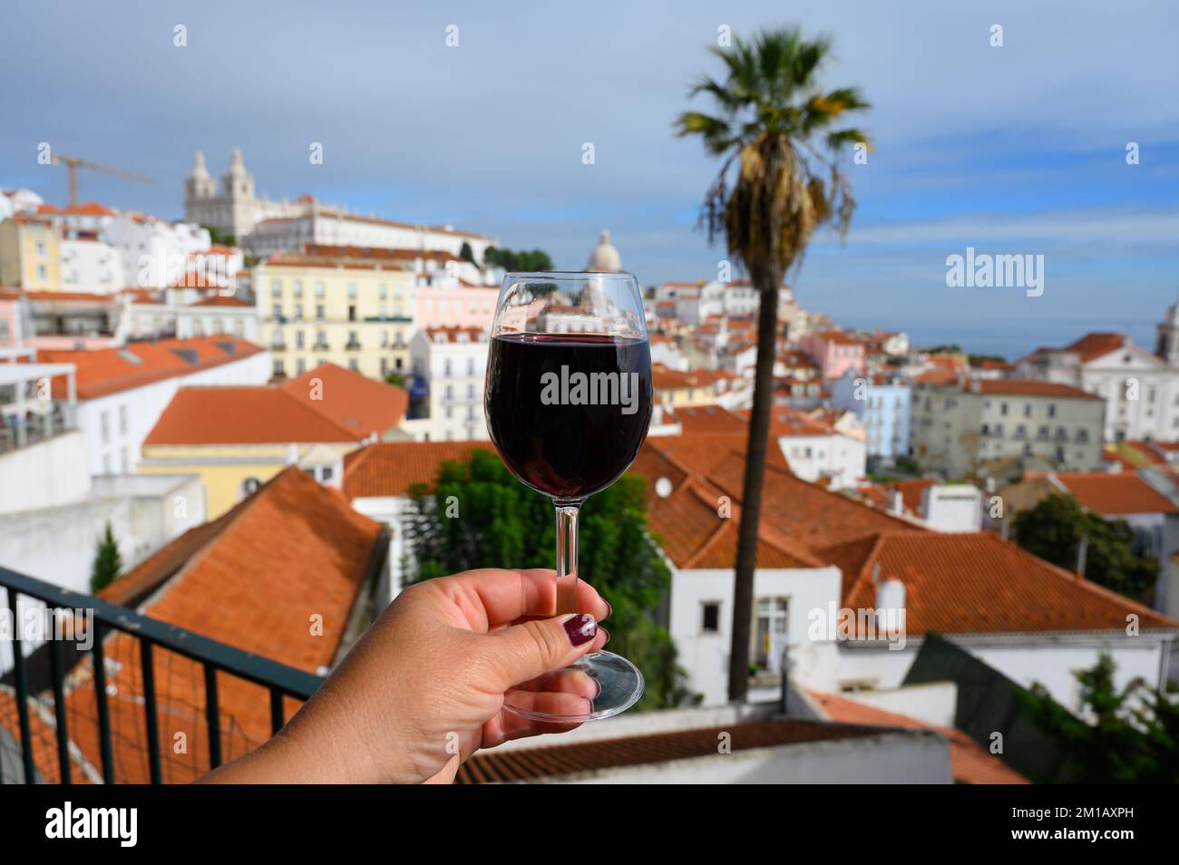 Hand with glass of dry red Portuguese wine in outdoor cafe at view point on colorful old part of Lisbon city, Portugal Stock Photo