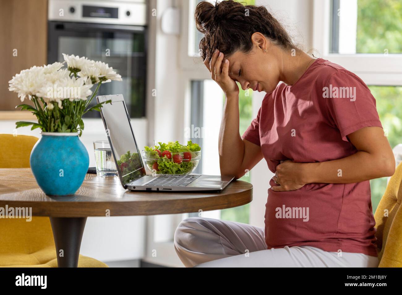 Pregnant woman sitting at table in front of laptop and feeling nauseous Stock Photo