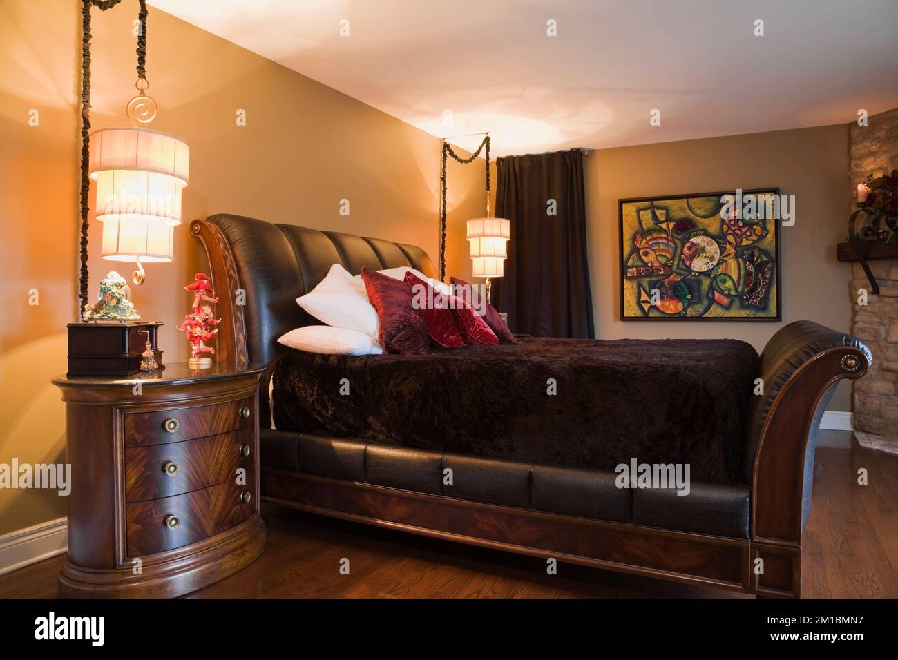 King size wood and leather sleigh bed and bedside table in master bedroom with tan yellow painted walls on upstairs floor inside luxurious home. Stock Photo
