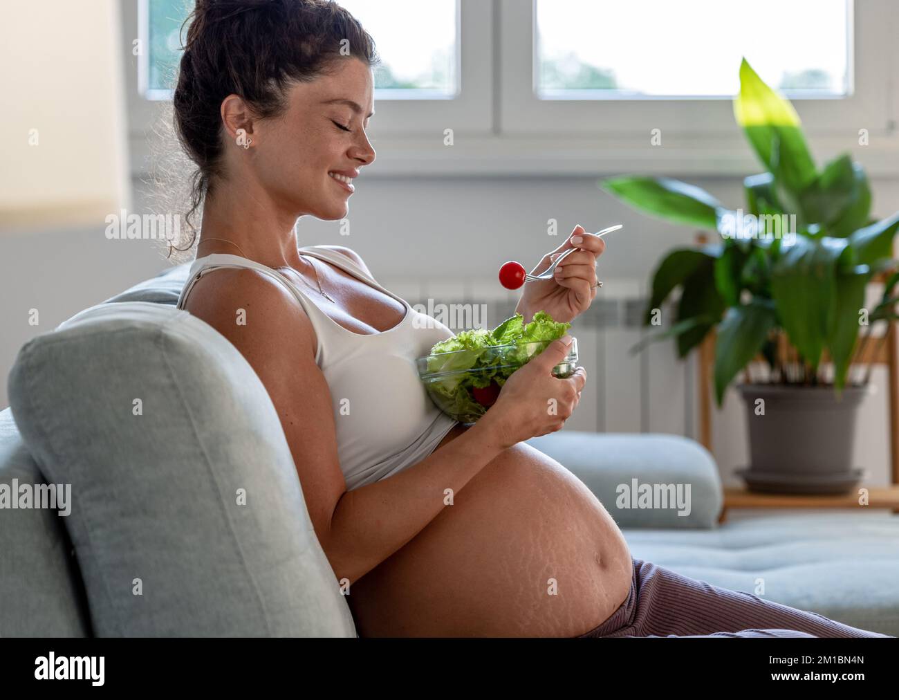 Pretty young woman eating salad on sofa at home. Healthy lifestyle during maternity period Stock Photo