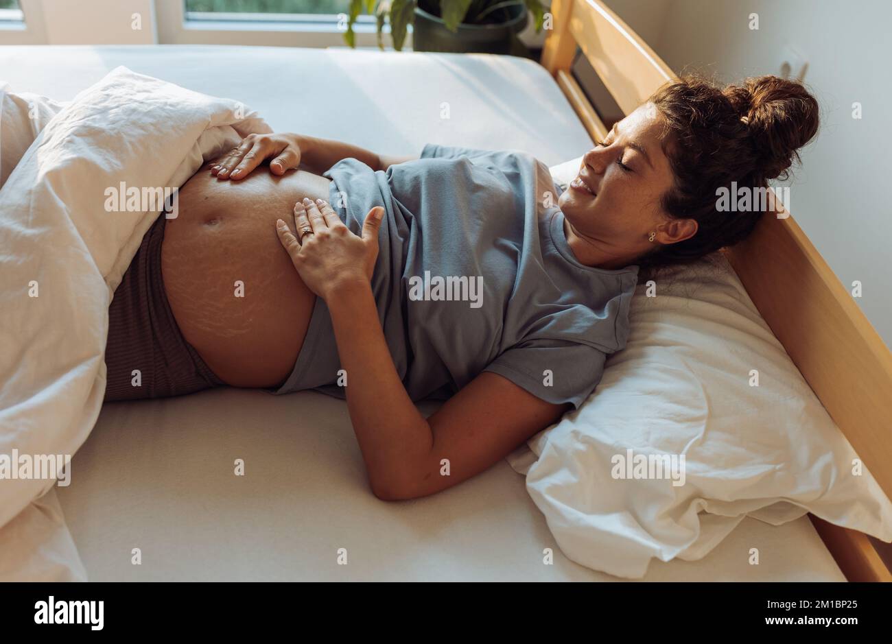 Pretty pregnant woman lying in bed, waking up, looking at belly and feeling happy Stock Photo