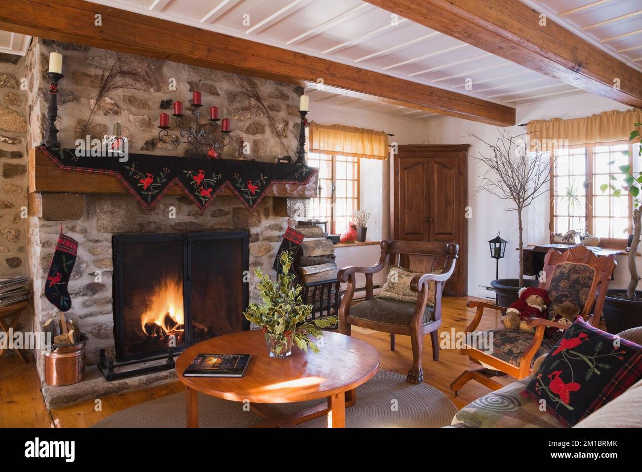 Lit fieldstone fireplace with antique wooden armchairs and Christmas decorations in living room inside old 1800s Canadiana cottage style log home. Stock Photo