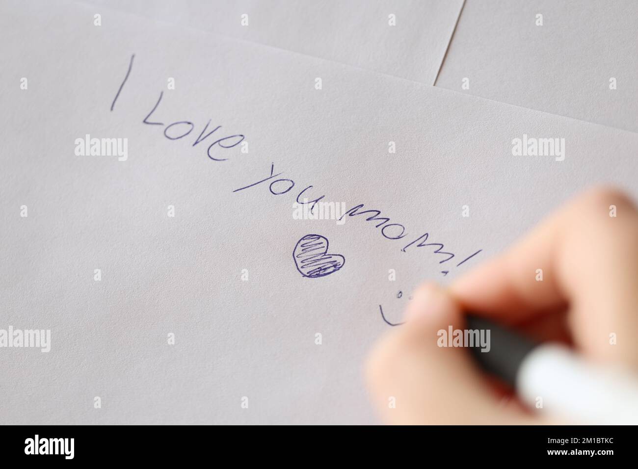Mothers day . I love you mom inscription on a white sheet and a heart and a childs hand with a pen. Stock Photo