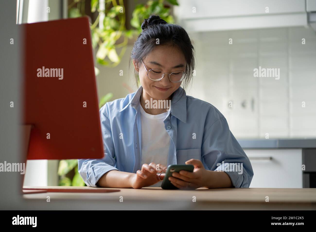 Smiling Asian girl freelancer sitting at home office using cellphone for personal during work hours Stock Photo