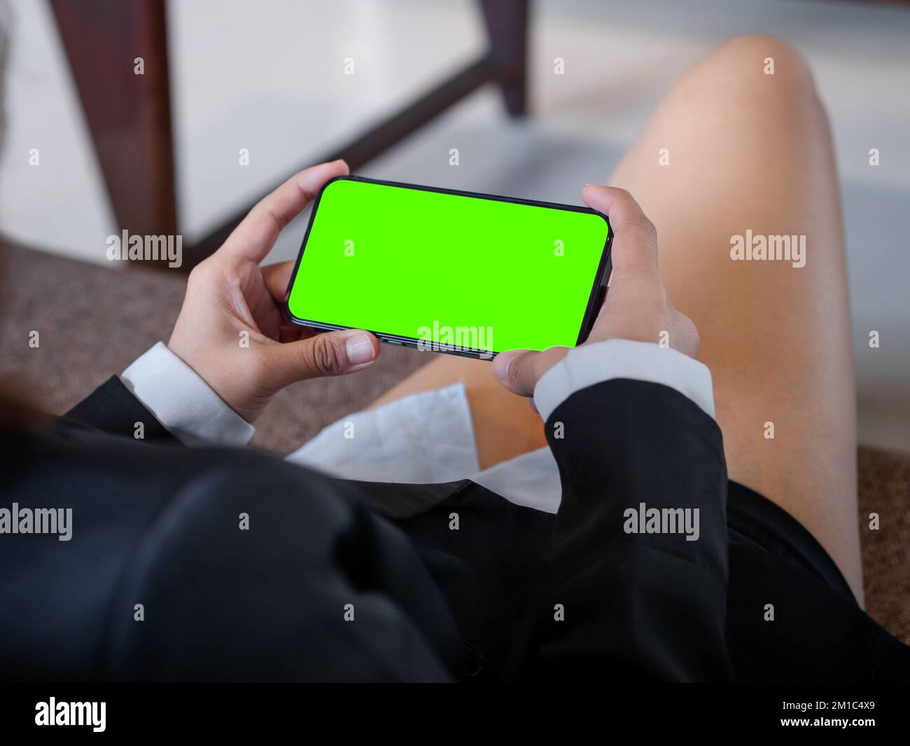 Green screen on mobile phone horizontal style in business woman's hands who wearing formal suit and shorts, relaxed sit with cross legs on couch in co Stock Photo