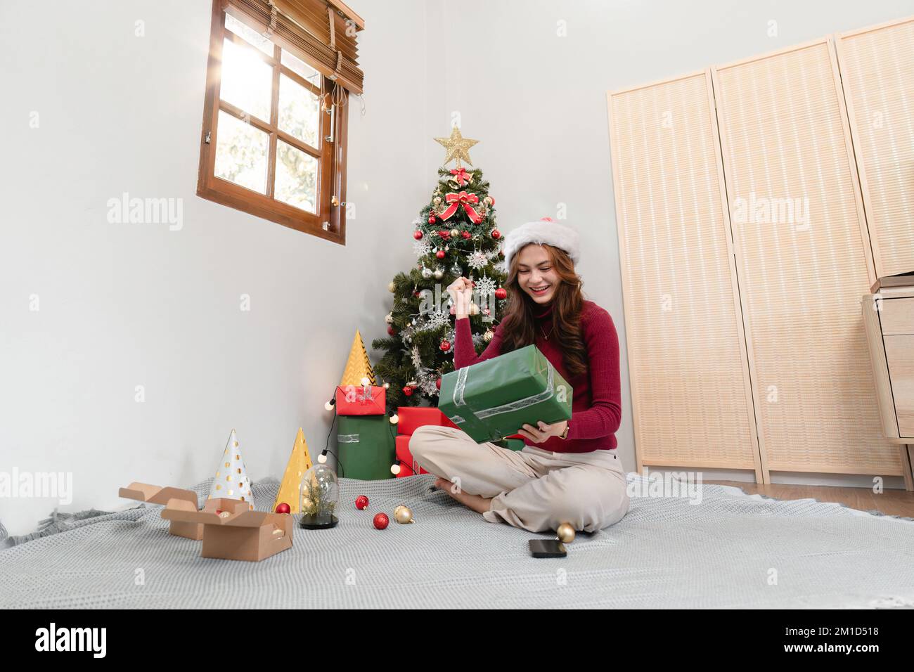 Happy woman sitting against Christmas tree background. Cheerful lady surprised of gift box. Marry Christmas and Happy Holidays Stock Photo