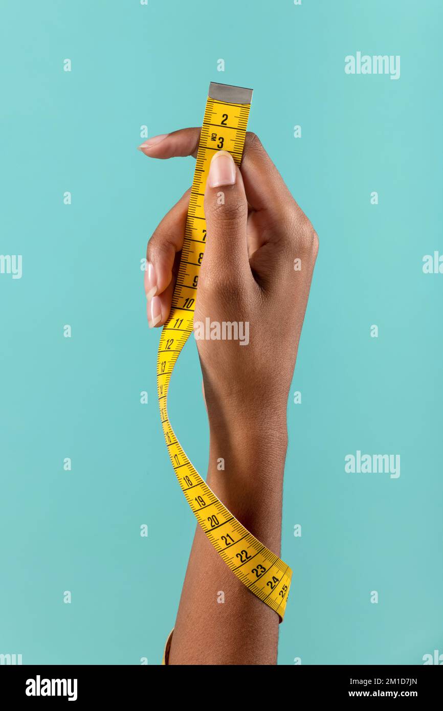 Crop anonymous African American female demonstrating yellow measuring tape wrapped around arm on turquoise background Stock Photo
