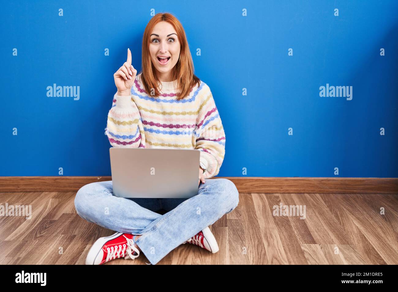 Young woman using laptop at home sitting on the floor pointing finger ...