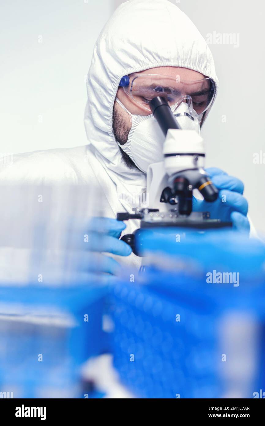 Man laborant in ppe suit using microscope doing research of coronavirus Scientist in protective suit sitting at workplace using modern medical technology during global epidemic. Stock Photo