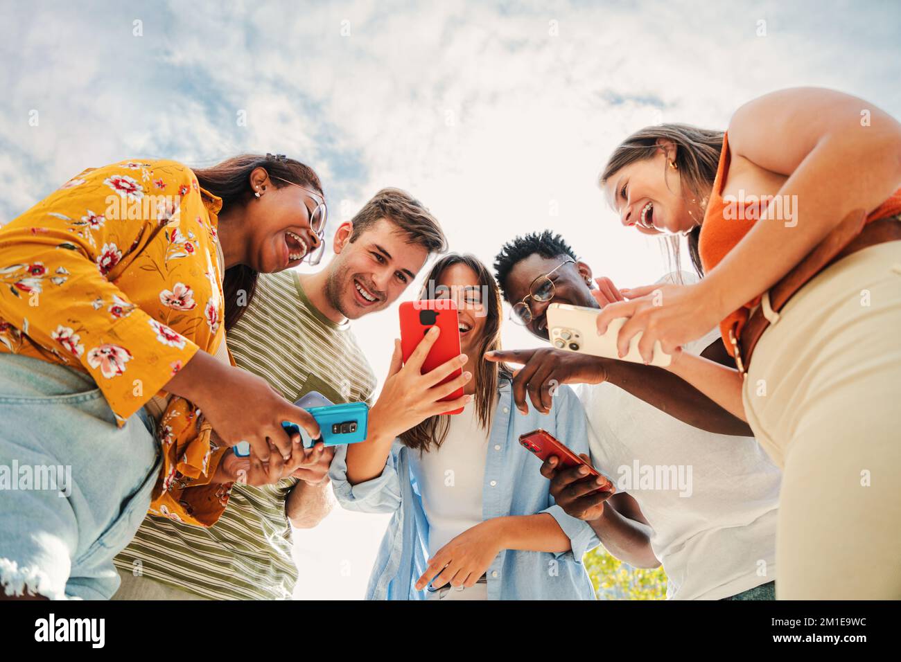 Low angle view of a group of smiling multiracial teenagers addicted to smartphones, watching funny videos, shopping online, enjoying outdoors. Multiethnic cheerful young people searching ,entertaining with different aplications on their mobile phones under blue sky. Connection concept. High quality photo Stock Photo