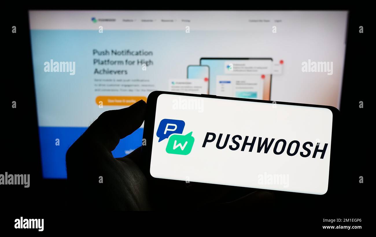 Person holding mobile phone with logo of American marketing platform company Pushwoosh Inc. on screen in front of webpage. Focus on phone display. Stock Photo