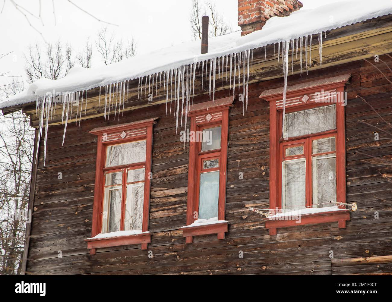 Icy, long icicles hang on the edge of the roof, winter or spring. Plank wall of an old wooden house with windows. Large cascades of icicles in smooth, beautiful rows. Cloudy winter day, soft light. Stock Photo
