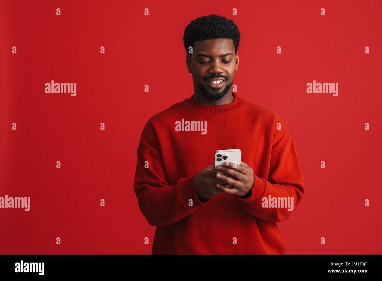 Black bearded young man smiling and using cellphone isolated over red background Stock Photo