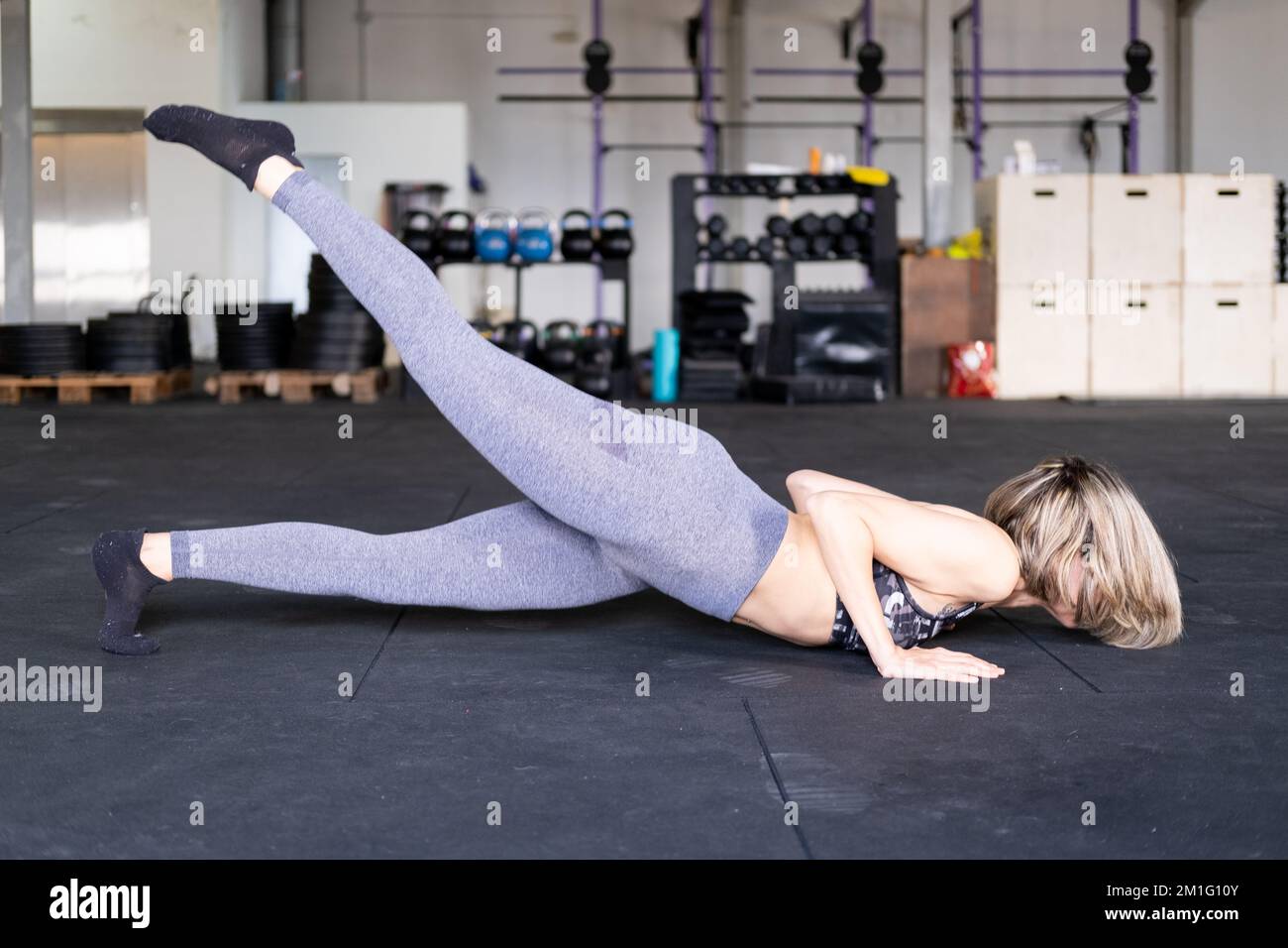An unrecognizable blond mid adult woman practicing vinyasa flow yoga with her right leg up and wearing leggings and a sports bra in a gym. Stock Photo