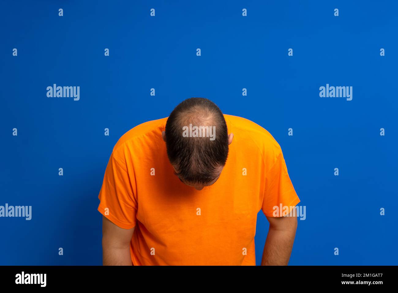 Man in an orange t-shirt leaning his head forward to reveal his bald spot, isolated on blue studio background. Stock Photo