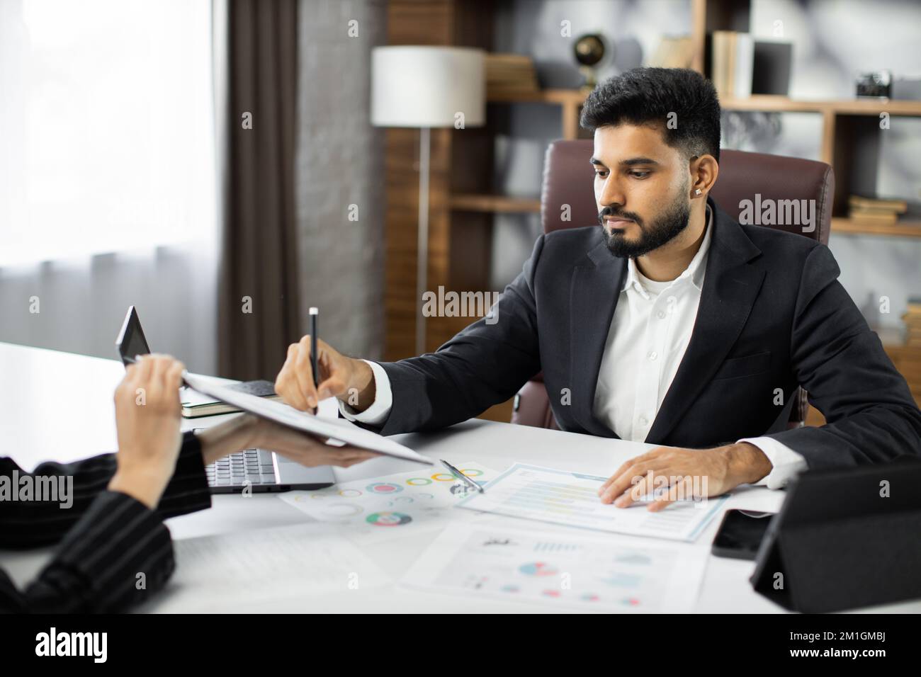 Focused young businessman signing agreement with skilled female lawyer. Concentrated financial advisor showing place for signature on paper contract document to male client at meeting in office. Stock Photo