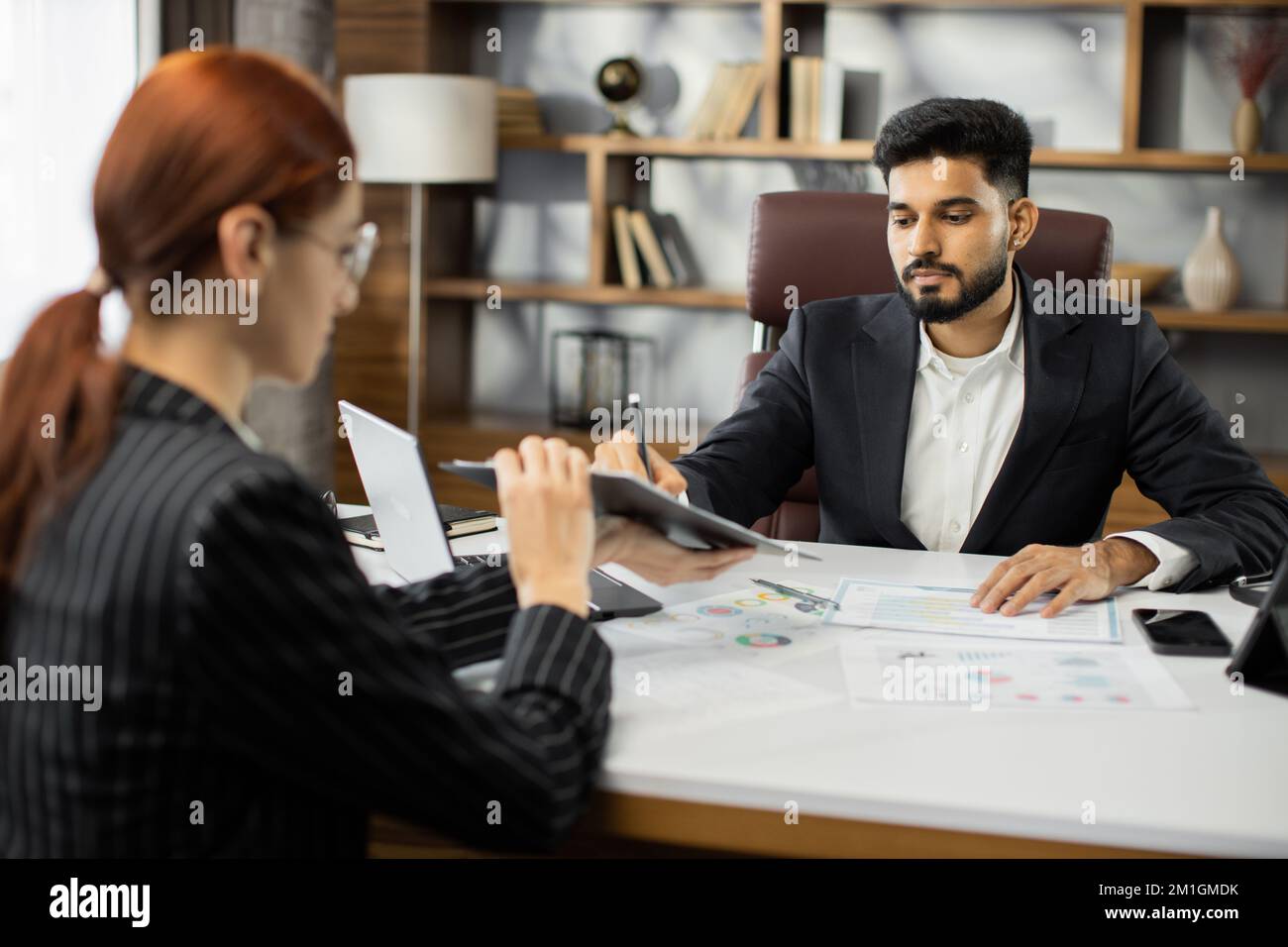 Focused young businessman signing agreement with skilled female lawyer. Concentrated financial advisor showing place for signature on paper contract document to male client at meeting in office. Stock Photo