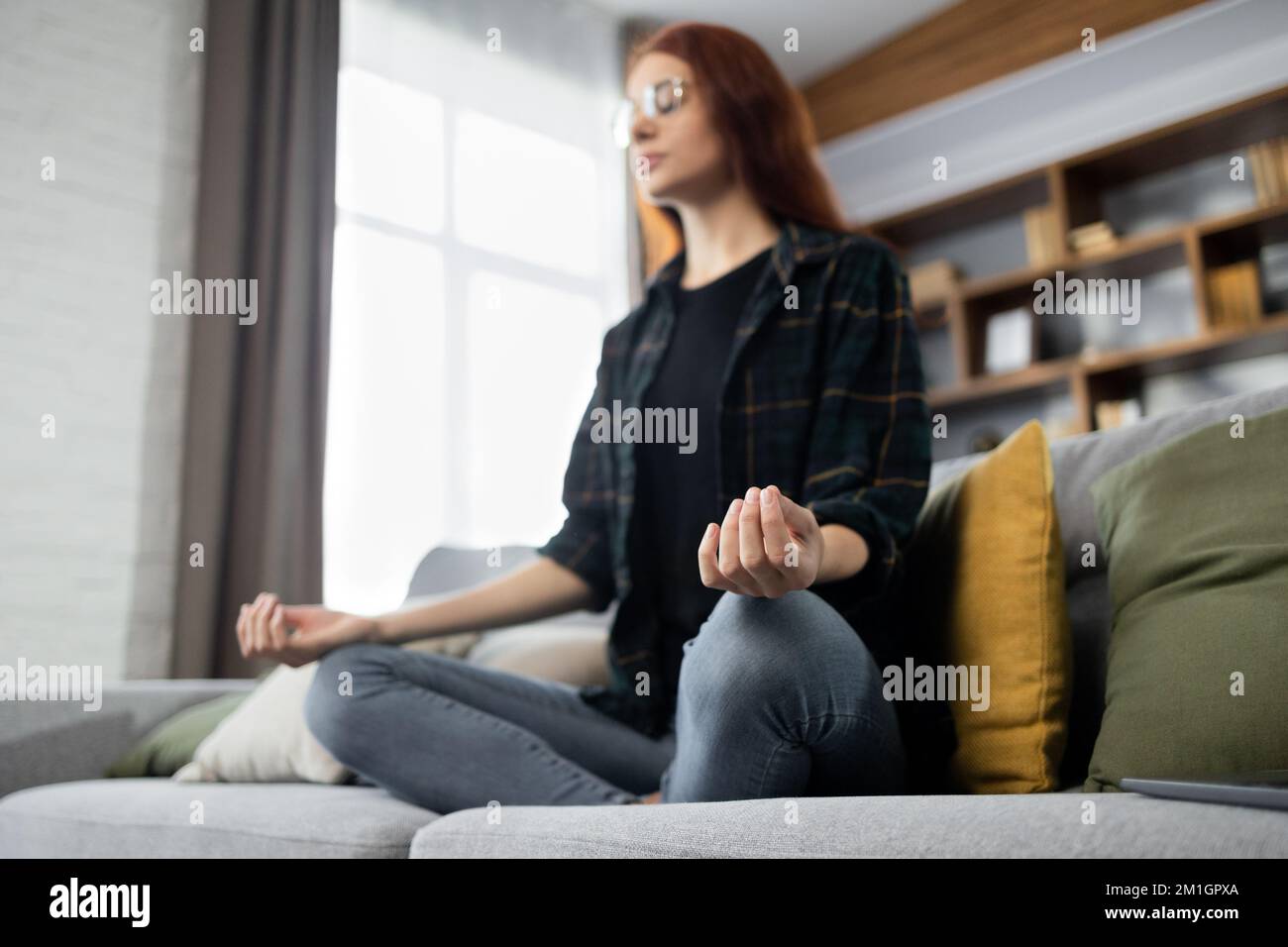 Yong female sitting on couch in lotus pose put hands on lap folded fingers closed eyes enjoy meditation do yoga exercise at home. No stress healthy habit mindfulness lifestyle anxiety relief concept Stock Photo