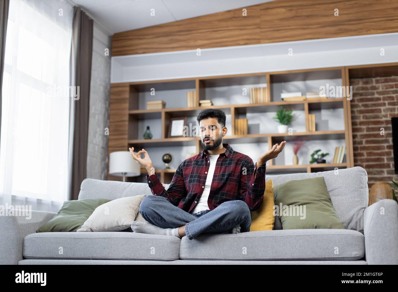 Yong male sitting on couch in lotus pose put hands on lap folded fingers closed eyes enjoy meditation do yoga exercise at home. No stress healthy habit mindfulness lifestyle anxiety relief concept Stock Photo