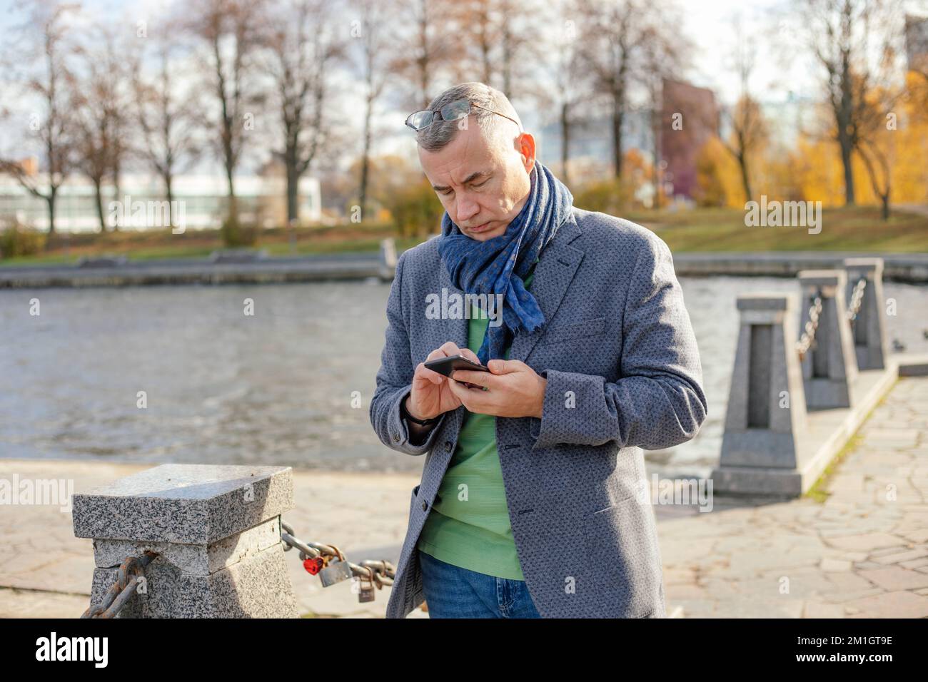 Portrait of middle-aged man standing near concrete pillars joined with chains in city in autumn, looking at smartphone. Stock Photo