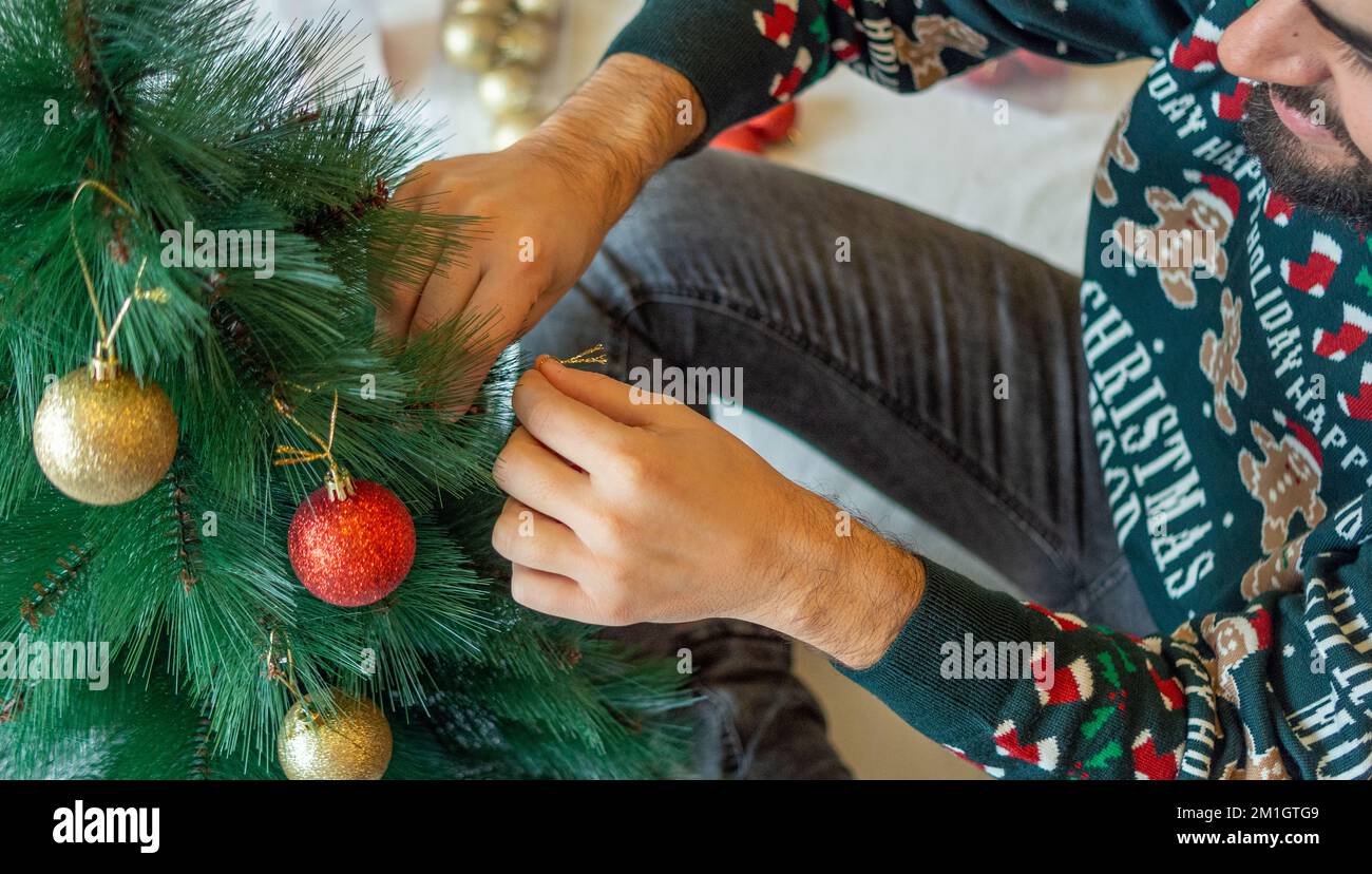 happy man putting a golden ball on the christmas tree wearing a christmas sweater Stock Photo