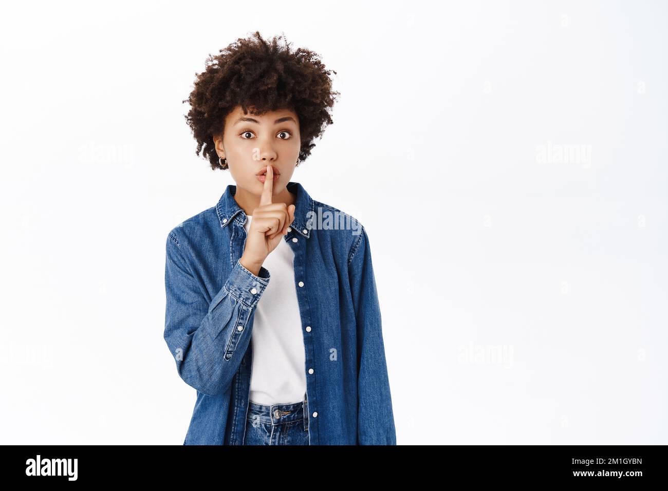 Portrait of Black woman shows hush, shhh shush gesture, taboo, asks to keep quiet, gossiping, stands over white background Stock Photo
